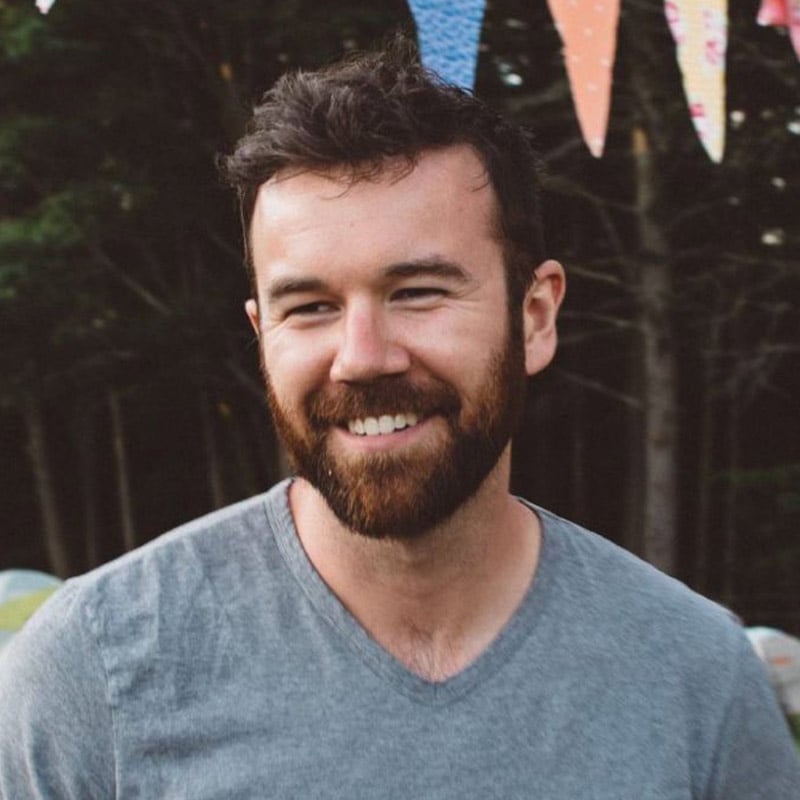 A bearded man with short dark hair smiles while standing outdoors. He is wearing a gray t-shirt, and colorful triangular flags are hanging above him. Trees are visible in the background.