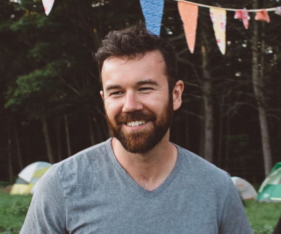 Bearded man in a gray t-shirt smiles outdoors, with colorful bunting overhead and tents in the background among trees—meet Rich Radimer, featured here for our Team Bio Popup.